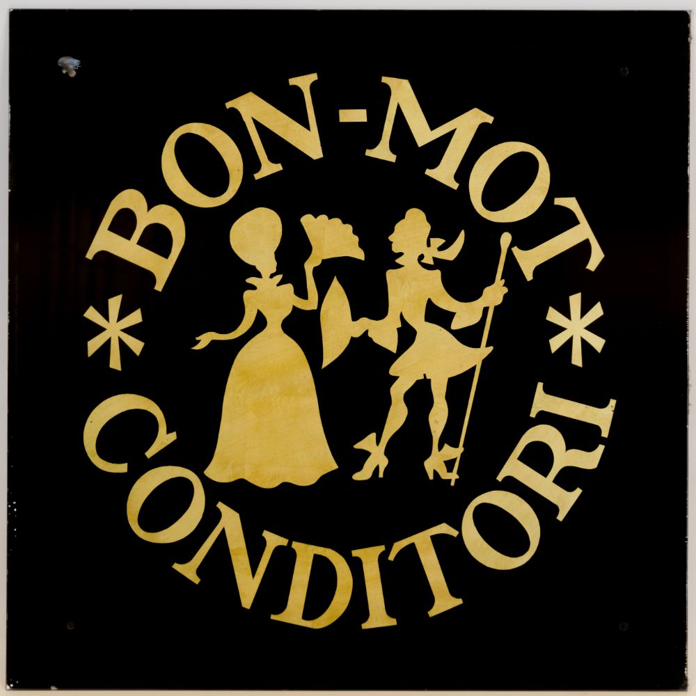 Svart skilt der det står med skrift i gull "Bon-Mot Conditori" og en stilisert kvinne og mann i pene klær. 
