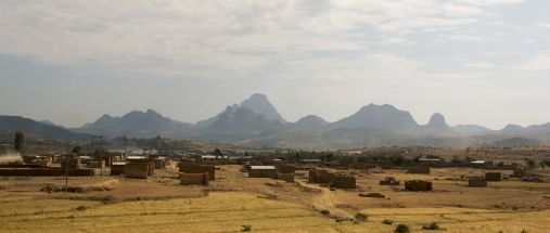 Houses on brown ground in front and tall mountains in the skyline.