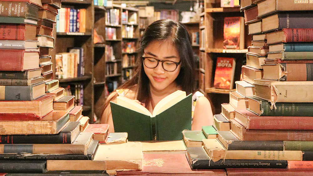 Ung jente med briller leser bok omgitt av store stabler med bøker.