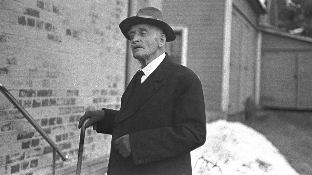 En eldre mann med hatt, frakk og stokk står i en trapp. Svart-hvitt-fotografi. 