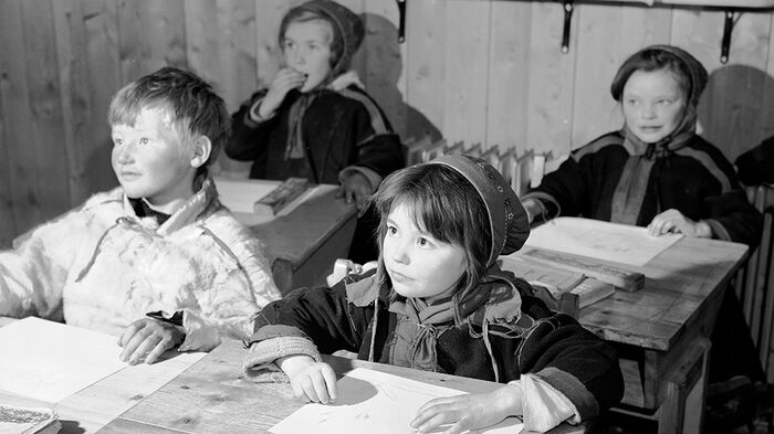 Four children sit at their individual school desks. They look straight ahead. The photo is in black and white.