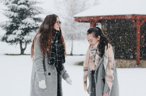 To unge kvinner står ute i snøværet og prater. Begge har langt, mørkt hår og er ikledd grå kåper, store skjerf og votter. 
