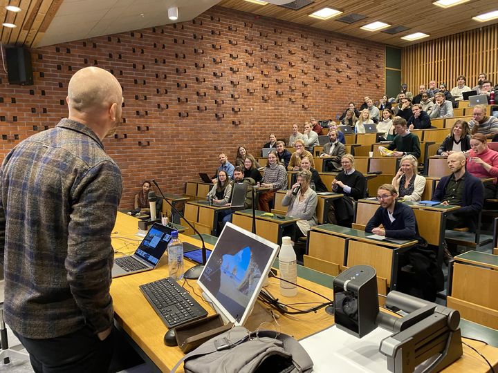 Et auditorium med mange mennesker og en mann som står framme og presenterer. Foto.