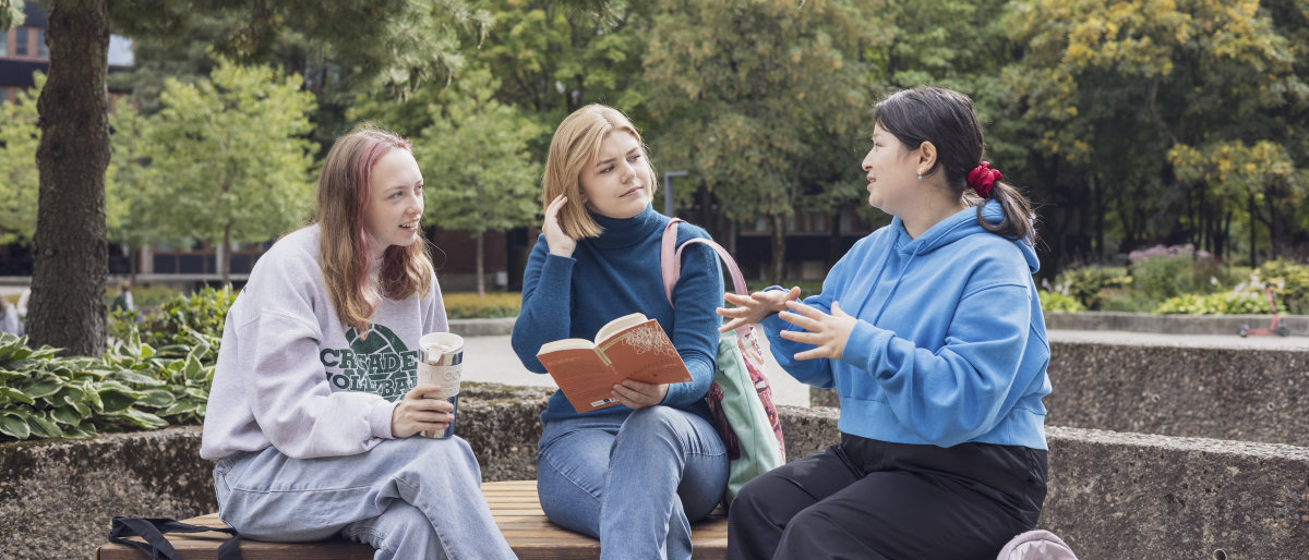 Tre studenter som sitter ute på campus og snakker og leser en bok. Foto.