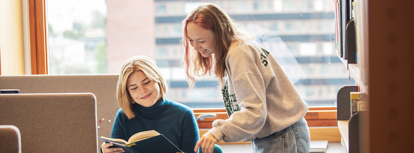 Two smiling students in the study hall, looking in a book together. Photo.