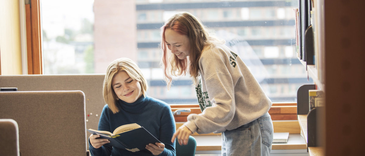 Two smiling students in the study hall, looking in a book together. Photo.