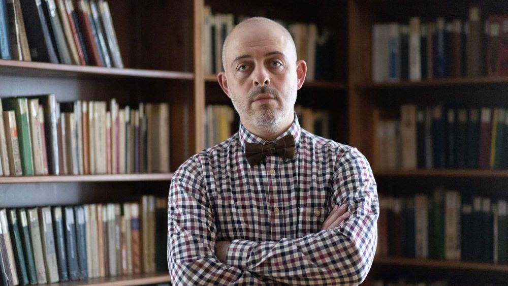 A man with no hair in a plaid shirt and bow tie. In the background there are several bookshelves full of books. Photo
