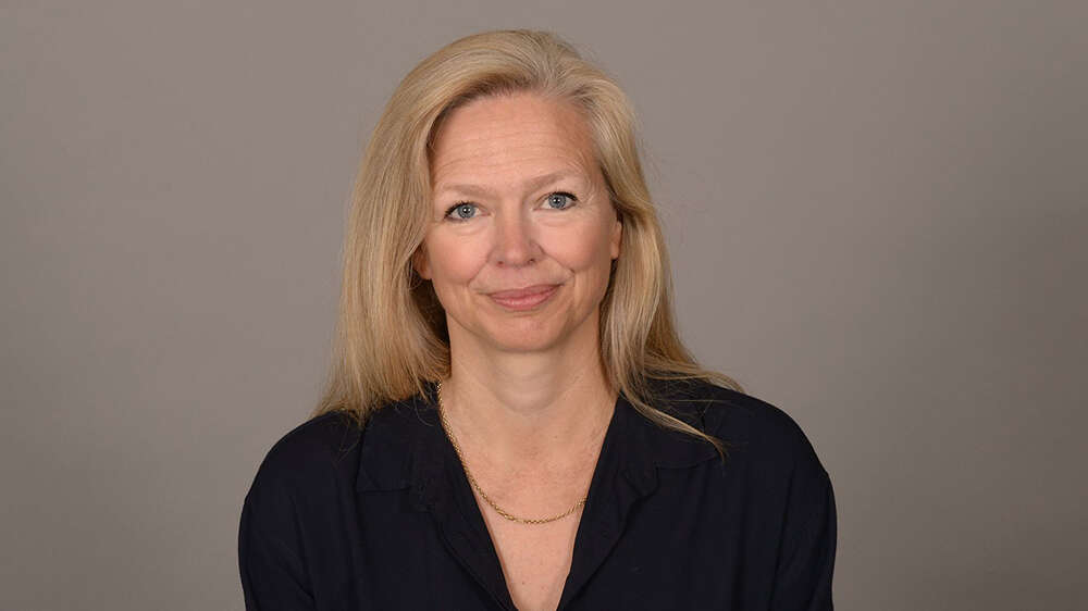 Portrett av en dame med lyst hår, sort overdel og grå bakgrunn.