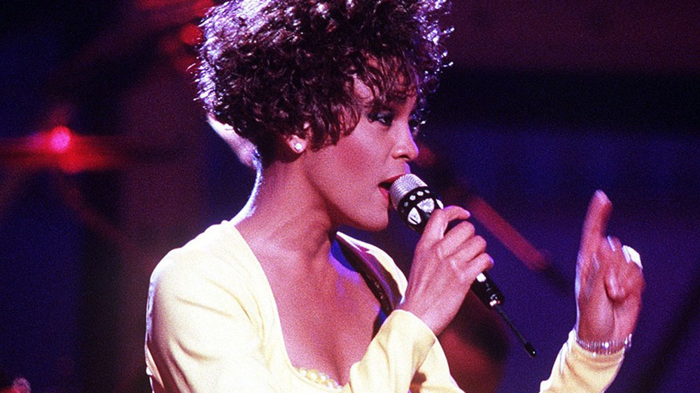 A woman with brown curly hair and a yellow top singing into a microphone. Photo.