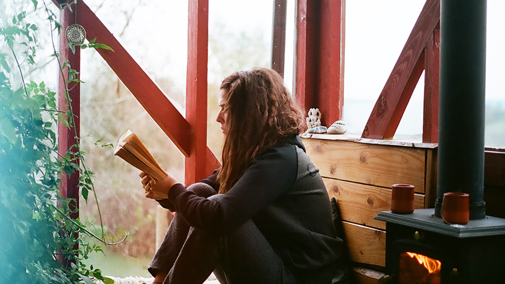 Kvinne sitter ved vindu og peis og leser i en bok. 