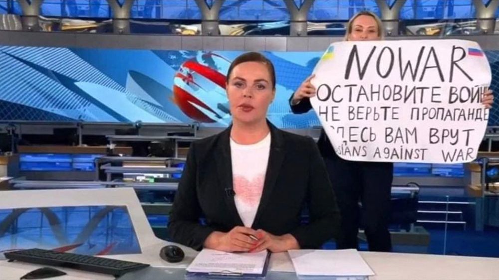 Et kvinnelig nyhetsanker i forgrunnen, i bakgrunnen holder en kvinne opp en plakat med blant annet ordene "no war". 