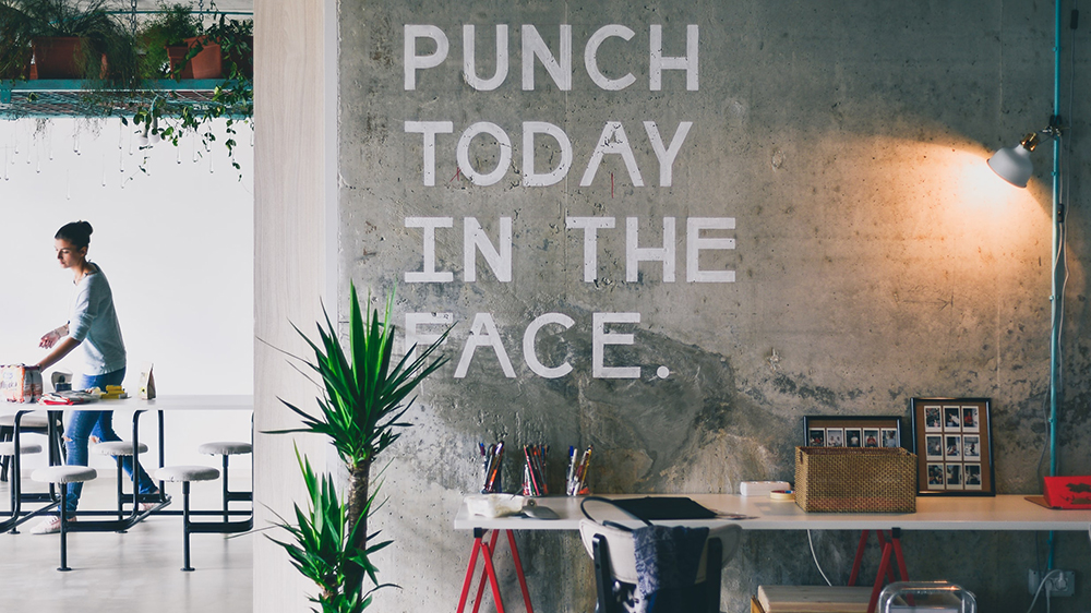 Et fotografi av et skrivebordshjørne med sementvegg og skriften "Punch today in the face" på veggen.