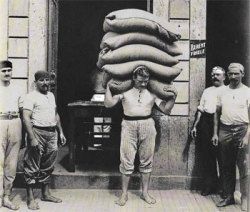 A man with lots of sand bags on his back. And other men standing besides him.
