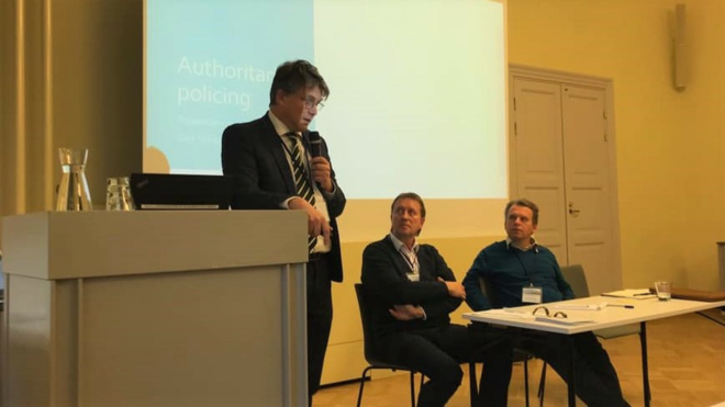 Professor Geir Flikke står i midten av bildet med en mikrofon og snakker ut til et publikum som er utenfor bildet. Bak ham er det en presentasjon. Til høyre for ham ser to kollegaer opp mens han snakker.