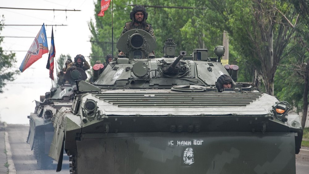 Tre stridsvogner kjører i kolonne igjennom en allé i Donbass. På toppen av stridsvognen står en soldat. På stridsvognen står det skrevet på russisk "Gud er med oss". Foto