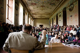 En mann med ryggen til rekker opp hånden og mang folk i en gammel hall. Foto.