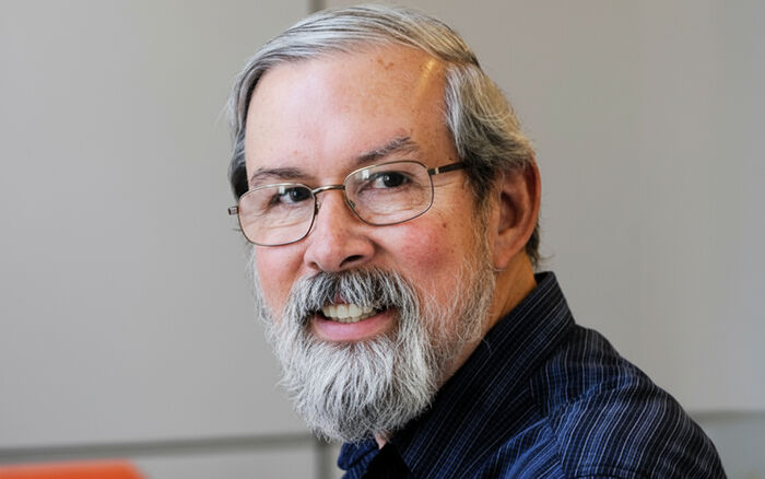 Portratit of a man with gray hair, grey beard, glasses and a dark shirt.
