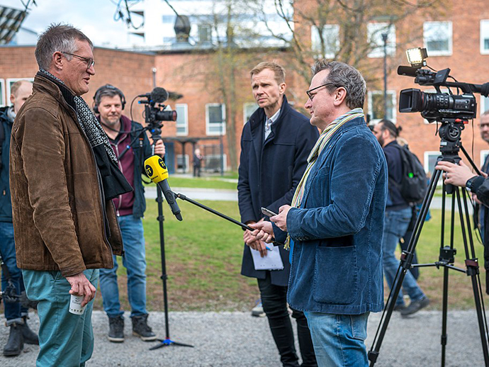 Anders Tegnell - tall man with grey hair wearing glasses and a brown leather jacket, surrounded by journalists, four men, some with camera or microphone.