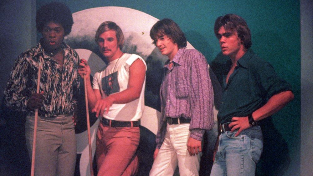 Bilde av fire unge menn, kledd i jeans og skjorter som lener seg inntil en vegg og holder biljardkø i hånden. Foto.