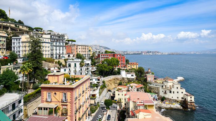 Bilde av et utsnitt av kysten i Napoli, Italia. Bygninger i bratt skrånng, havn, vann, hav, biler, pent vær.