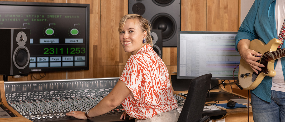 Students in the music studio, one is sitting by the mixing table in front of a screen and one is standing with a guitar. Photo.