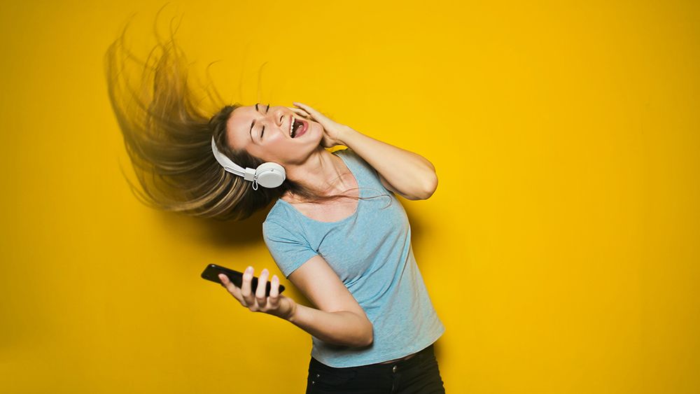 Blond, bright-skinned woman wearing a blue t shirt dances and sings wearing a white headset and holding a smart phone in her right hand. Yellow background. 