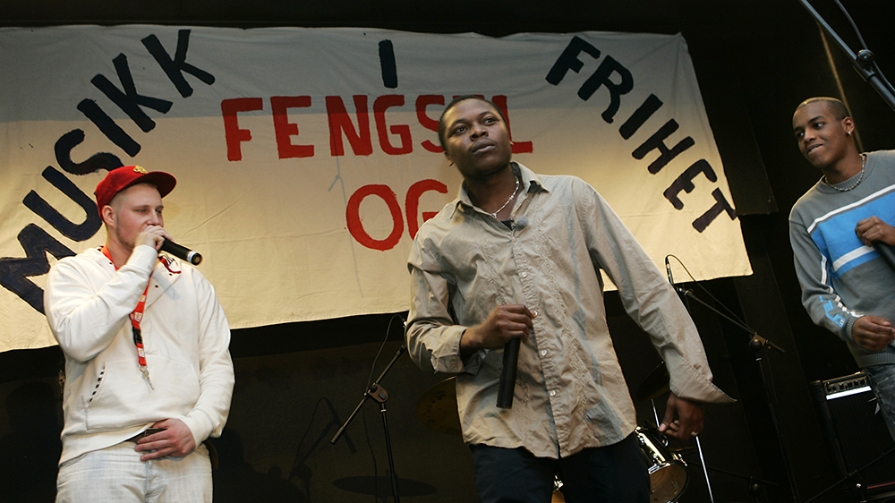 Three men on stage in front of a banner saying "Music in prison and freedom" in Norwegian.