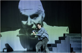 Scenebilde: skuespiller i dramatisk positur foran projeksjon av mannsansikt. Blå og grønne farger. Fotografi.