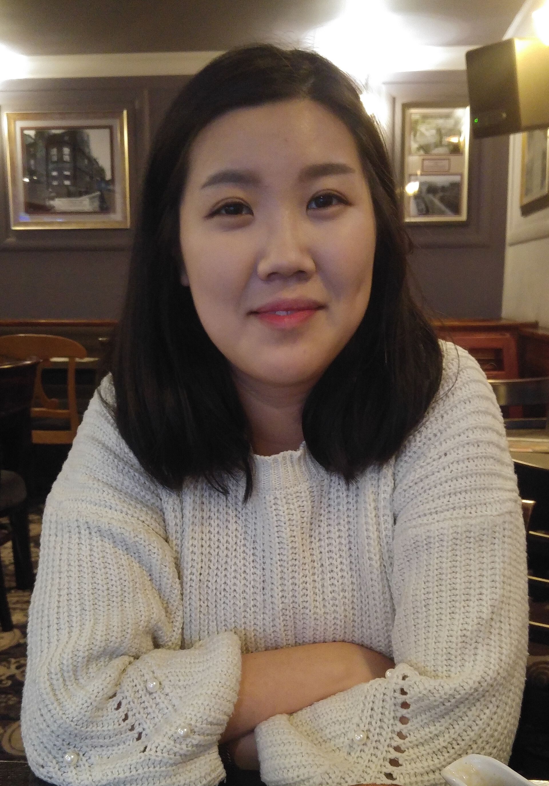 Bilde av Camilla Cho, en ung kvinne med lys hud og halvlangt, mørkt, rett hår, ikledd hvit genser og med armene i kors. Bildet er tatt innendørs ved et bord.