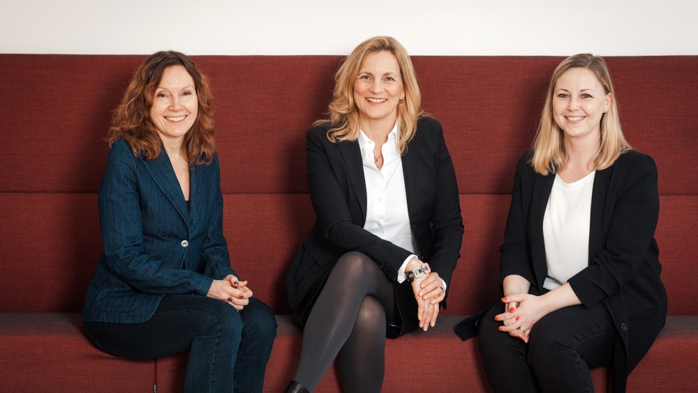 Ingebjørg Tonne, Unn Røyneland og Anne Charlotte Lindblom-Rosén sitter smilende på en rød sofabenk