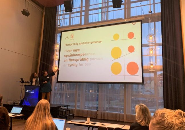Pernille Hansen og Elisabeth Holm  (ved Oslomet) presenterer på konferansen MONS 18. Dei står til ventre i biletet, og Hansen peiker på lysbileteframvisinga. På lysbiletet står følgjande tekst: "Hvor mye av språkkompetansen til en flerspråklig person er synlig for oss?". Lysbiletet viser òg ein figur som illustrerer balansen eller ikkje-balansen i fleirspråkliegheiten til ein person.