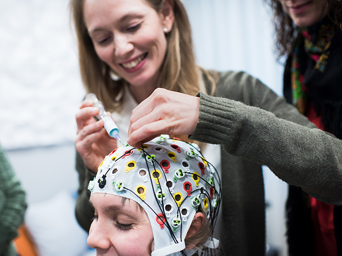 A woman wearing an EEG cap, another woman putting it on. Photo.