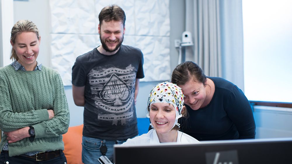 En kvinne med EEG hette sitter foran en skjerm, flere personer står rundt. Foto.