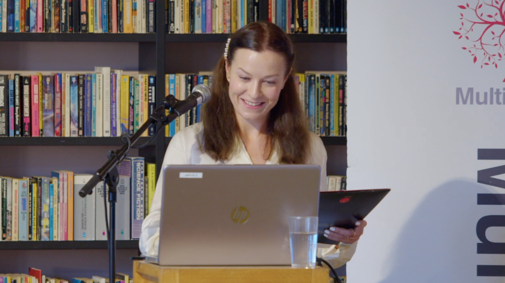 Oliwia Szymanska presenting, smiling, computer, microphone, book case 
