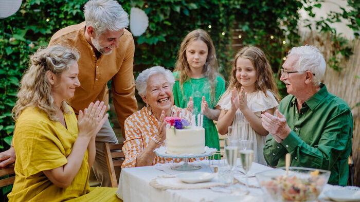 Picture of a family/birthday party with five family members clapping and gathering around a woman seated in front of a large cake. 