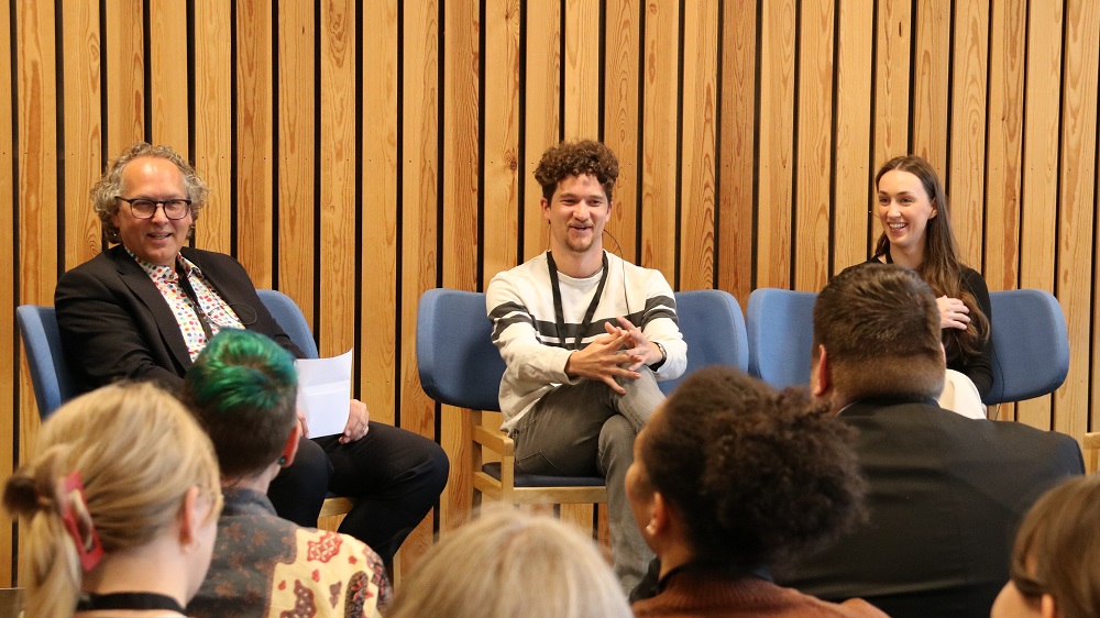 En mann og to studenter sitter på en scene. De smiler og ler. Fotografi. 