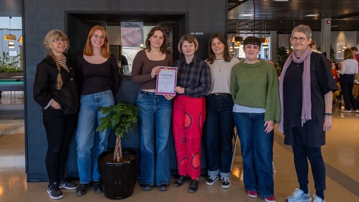 Bilde av en gjeng studenter som står sammen med fakultetsledelse inne i en kantine, og har et diplom i hånden. 