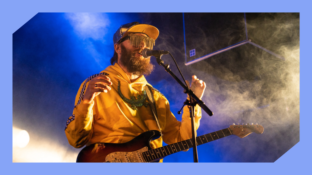 Mann med caps, solbriller og gitar står med en mikrofon foran seg. Det er skarpt scenebelysning og røyk i luften. Foto.