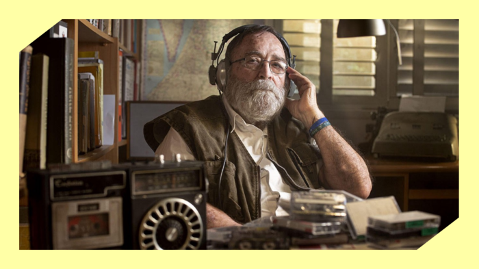 Foto av en eldre mann med hvitt skjegg og briller som sitter på et kontor og hører på et opptak på øreklokker mens han ser inn i kameraet.