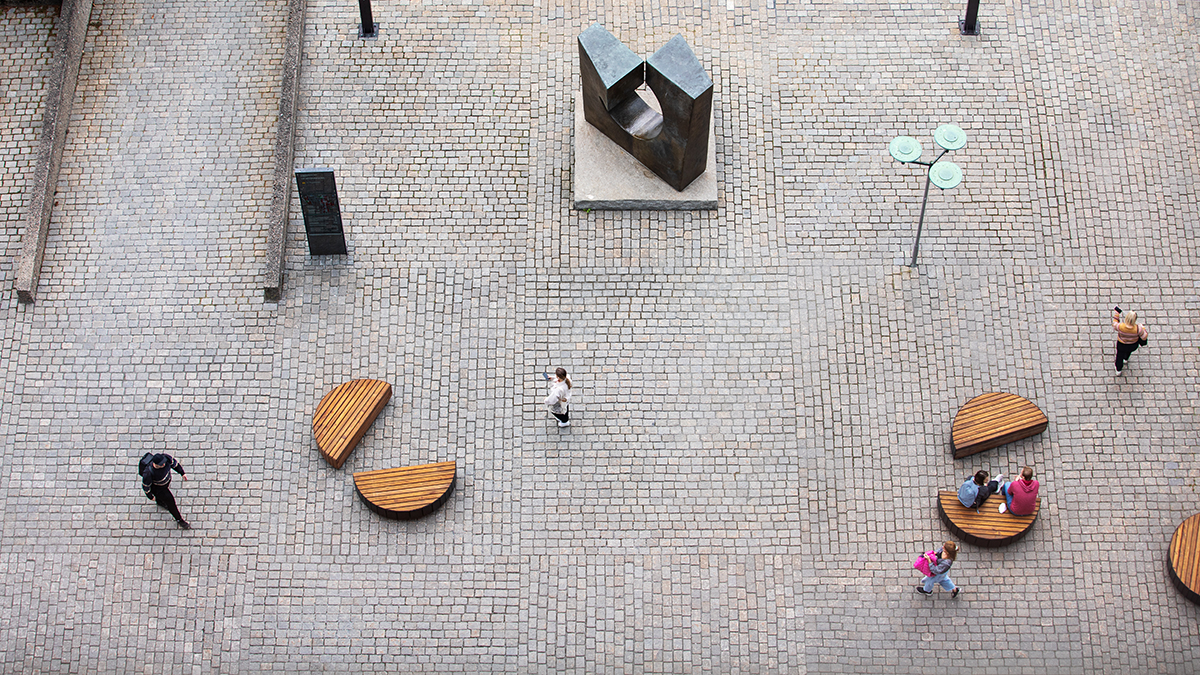 The area in front of Niels Treschows hus seen from above. People walking across the area, benches, lampposts and a sculpture. Photo.
