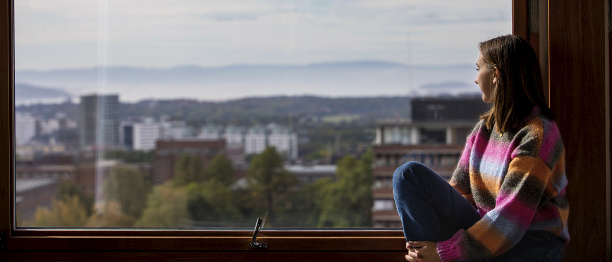 A student sitting on the windowsill looking out over the campus and Oslo. Photo.
