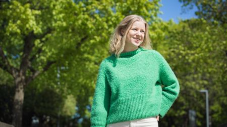 Smilende ung kvinne med grønn genser med trær i bakgrunnen. Foto