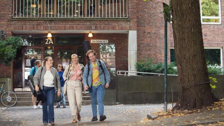 Three students with musical instruments in front of brick building. Photo