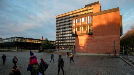Folk som krysser en plass foran en bygning i solnedgang. Foto 