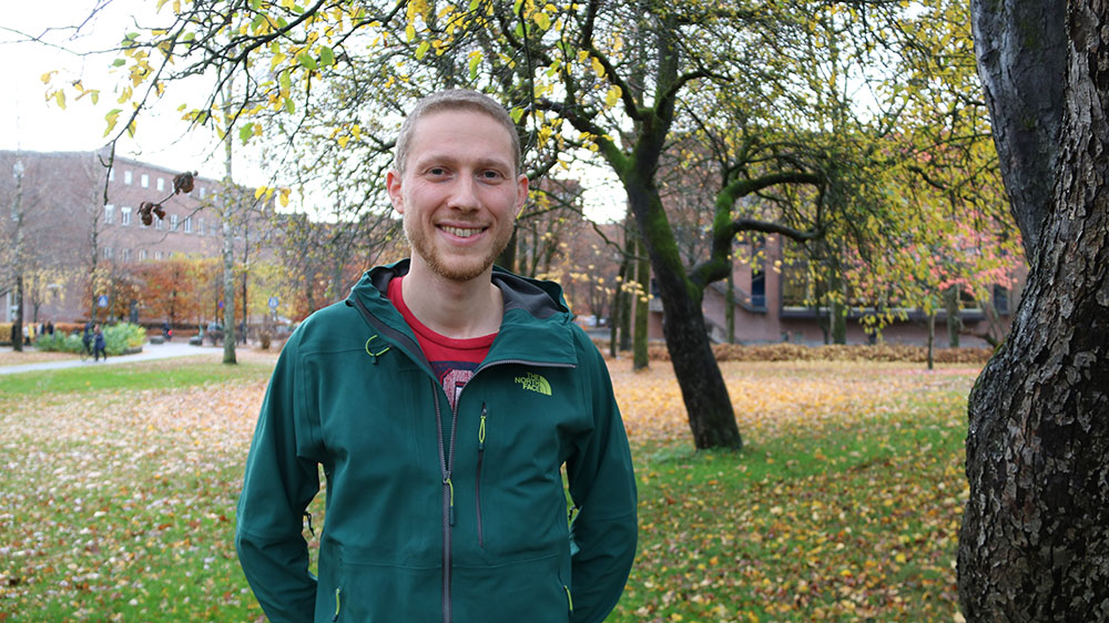 smilende mann i grønn anorakk blant trær på Blindern Campus