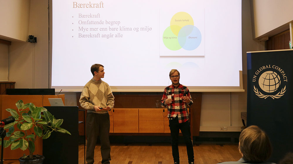 Foto av to studenter som står foran en forsamling i et auditorium.
