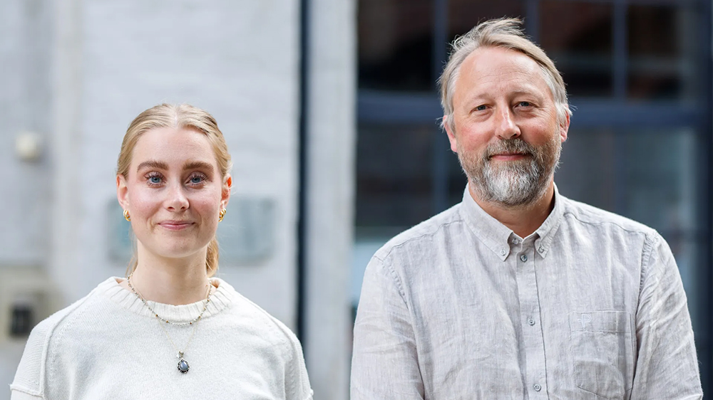 Bildet inneholder en ung kvinne med lyst hår og en middelaldrende mann med skjegg og grå grå skjorte. De står foran bygningen til norsk institutt for fredsforskning.