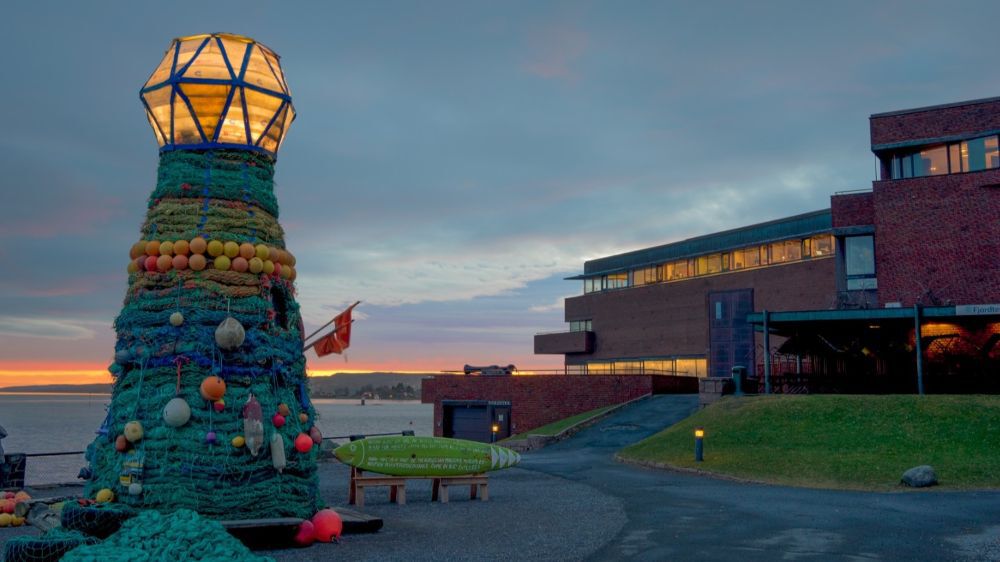 Et cirka fem meter høyt fyrtårn tildekket av fiskegarn i blå-grønne toner, samt blåsebøyer og fendere i ulike farger. I bakgrunnen ser vi havet og en mursteinsbygning på fire etasjer.
