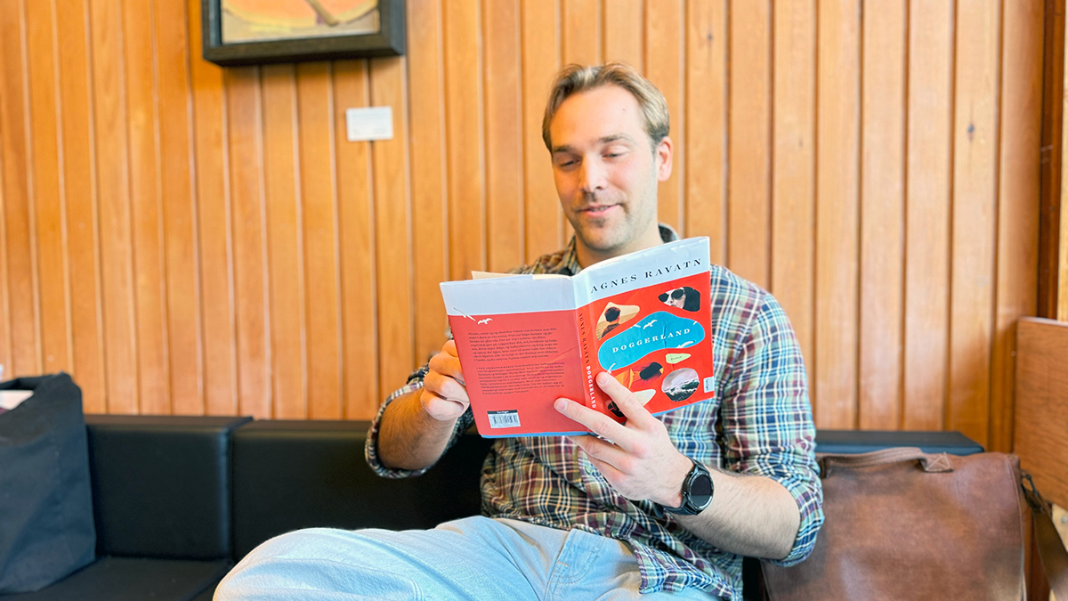 Foto av en ung mann med blondt som sitter og leser en bok ved et bord.