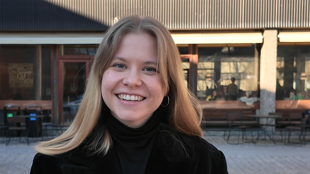 En ung kvinne ser rett frem. I bakgrunnen vises en vegg med vinduer. Foto. 